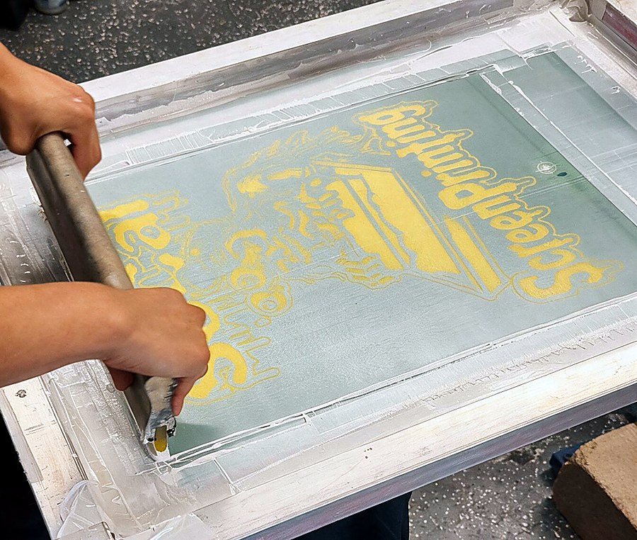 Hands pulling a squeegee loaded with yellow ink across a screen